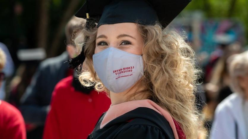 A graduate student looks back the camera and smiles while wearing a mask outdoors.